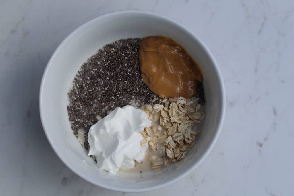 Healthy Snickers Overnight Oats (With Yogurt) White bowl full of ingredients needed to make Healthy Snickers Overnight Oats. A bowl of yogurt, chia seeds, oats, almond milk, peanut butter
